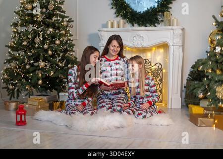 La mattina di Natale leggono insieme la famiglia. Una famiglia che indossa pigiami natalizi abbinati ama leggere un libro insieme vicino al caminetto, circondato Foto Stock