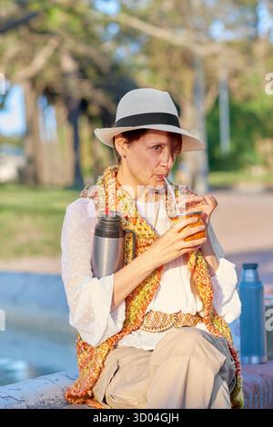 Una donna assaggia una tradizionale bevanda yerba mate in un parco all'aperto, vestito in stile artigianale bohémien. Foto Stock