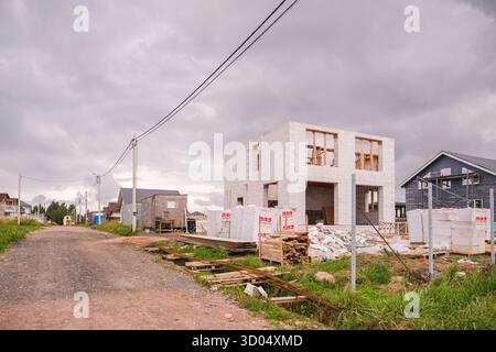 Russia, San Pietroburgo, 23.08.2025 costruzione incompiuta di una casa bianca con finestre aperte accanto a una strada sterrata e linee elettriche su un'overca Foto Stock