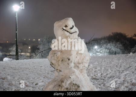 Pupazzo di neve fatto a mano con le braccia del bastone e il viso in piedi su una collina innevata di notte sotto il bagliore dei lampioni Foto Stock