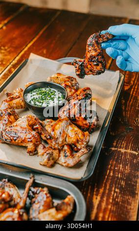 Una persona sta tenendo in mano un pezzo di pollo con un guanto blu. Il pollo si trova su un vassoio con una salsa Foto Stock