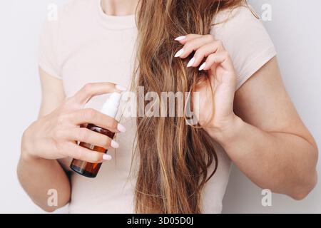Donna che spruzza la nebbia dei capelli. Routine per capelli castani lunghi. Applicare il liquido in balsamo. Trattamento di bellezza a casa. Primo piano mani che tengono la bottiglia. Foto Stock