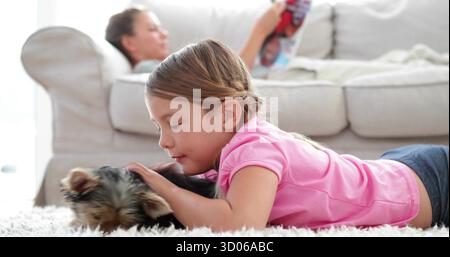 Accarezzare lo Yorkshire Terrier, ragazza che indossa una camicia rosa sdraiata su un tappeto bianco nel soggiorno di casa Foto Stock