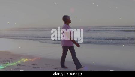 Donna anziana che cammina indossando T-shirt rosa e pantaloni grigi sulla costa sabbiosa, con cielo pastello Foto Stock
