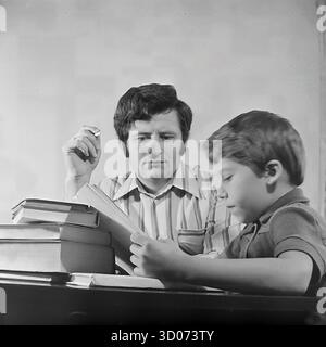 Un padre sovietico, un macchinista, guarda attentamente suo figlio leggere un libro a casa a Sloviansk, 1970-1980 Questa foto d'archivio in bianco e nero dall'URSS, la RSS Ucraina, è un ritratto intimo di padre e figlio. L'uomo, con una camicia a righe, tiene una sigaretta mentre osserva il ragazzo, che è concentrato sui suoi studi. Una grande pila di libri si trova sul tavolo lucido. Questa immagine cattura una scena della vita quotidiana sovietica, della paternità, dei valori familiari e dell'istruzione a casa. Un documento nostalgico di vita pacifica nel Donbas prima della guerra. Foto Stock