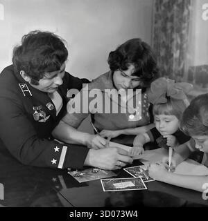Una famiglia sovietica felice di quattro persone (padre, madre, figlio e figlia) sta scrivendo biglietti d'auguri natalizi a casa a Sloviansk, 1970-1980 Foto d'archivio in bianco e nero, URSS, RSS Ucraina. Il padre, un macchinista e veterano, indossa la sua uniforme ferroviaria formale con medaglie. Tutta la famiglia si siede insieme al tavolo. Anche i bambini scrivono. Questa coinvolgente immagine cattura una scena della vita quotidiana sovietica, delle tradizioni familiari e dei genitori. Un documento nostalgico di una famiglia felice e di una vita pacifica nel Donbas prima della guerra. Foto Stock