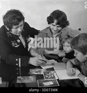 Una famiglia sovietica felice di quattro persone (padre, madre, figlio e figlia) sta scrivendo biglietti d'auguri natalizi a casa a Sloviansk, 1970-1980 Foto d'archivio in bianco e nero, URSS, RSS Ucraina. Il padre, un macchinista e veterano, indossa la sua uniforme ferroviaria formale con medaglie. Tutta la famiglia si siede insieme al tavolo. Anche i bambini scrivono. Questa coinvolgente immagine cattura una scena della vita quotidiana sovietica, delle tradizioni familiari e dei genitori. Un documento nostalgico di una famiglia felice e di una vita pacifica nel Donbas prima della guerra. Foto Stock