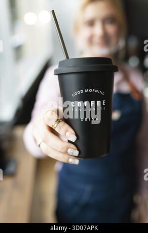 Barista ragazza che tiene in mano un bicchiere di metallo nero con l'iscrizione buon mattino preferito caffè drink in mano con caffè caldo preparato. Professionale c Foto Stock