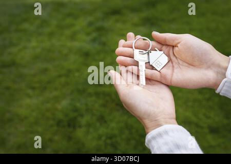 Chiavi con un portachiavi in forma di casa di metallo da una nuova casa o appartamento nelle mani di una ragazza su uno sfondo di erba verde Foto Stock