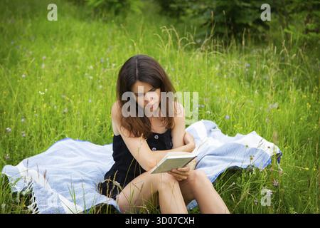 Picnic all'aperto. Una ragazza legge un libro all'aperto mentre si siede su una plaid blu. La ragazza gode dell'aria fresca. Attività ricreative all'aperto, primo piano Foto Stock