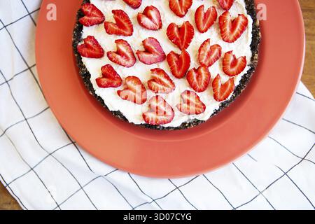 Cheesecake alla fragola decorata con fragole a forma di cuore giace su un piatto color corallo, si erge su un tovagliolo e un tavolo di legno. Immagine di una Foto Stock