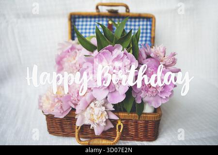 Un bellissimo bouquet di peonie rosa in un vaso si erge su una valigia di legno. Bella composizione. Iscrizione buon compleanno Foto Stock