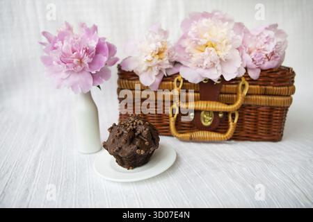 Muffin al cioccolato e bellissime peonie rosa giacciono su una valigia di legno. Bella composizione Foto Stock