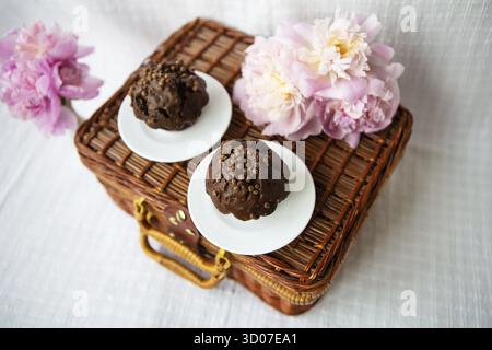 Muffin al cioccolato e bellissime peonie rosa giacciono su una valigia di legno. Bella composizione Foto Stock