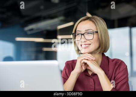 Donna d'affari felice che indossa occhiali da vista e una camicia marrone, sorridendo allegramente mentre lavora su un notebook in un moderno ufficio aziendale Foto Stock