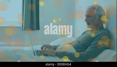 Un anziano che scrive su un computer portatile in pigiama sul letto con lampada, cuscini, tende della finestra, spazio per fotocopie Foto Stock