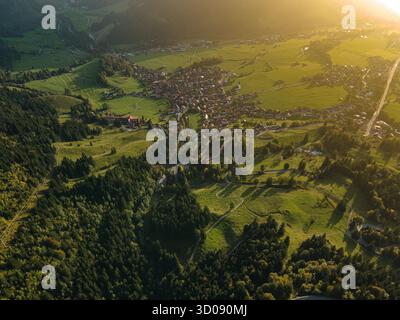 Vista aerea di prati verdi lussureggianti che incontrano fitte foreste, un pittoresco villaggio immerso nel mezzo, immerso nel caldo bagliore del sole che tramonta, Bad Hindelang, Bayern, Deutschland. Foto Stock