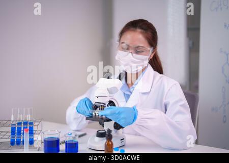 Una donna asiatica scienziata adulta esegue diligenti analisi microscopiche in un laboratorio sterile indossa attrezzature protettive mentre esplora la scienza Foto Stock