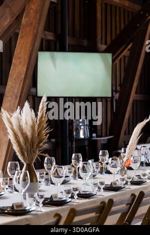 Un elegante tavolo da nozze all'interno di un fienile rustico presenta biancheria bianca, piatti neri, bicchieri, pampas in erba e uno schermo di proiezione Foto Stock