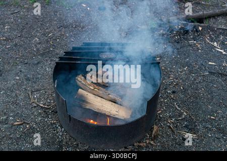 Una fossa antincendio di metallo si illumina di tronchi in fiamme, inviando segnali di fumo verso il cielo. Takakkaw Falls, British Columbia Foto Stock