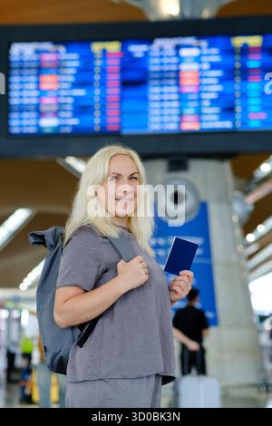 Una donna si trova in sicurezza presso un terminal dell'aeroporto, con un passaporto e uno zaino, mentre guarda verso il bordo di partenza del volo e othe Foto Stock