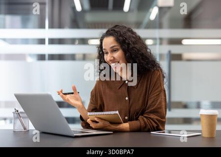 Giovane donna felice che partecipa attivamente a una videochiamata online su un notebook di un ufficio moderno, prendendo appunti e discutendo concetti aziendali Foto Stock