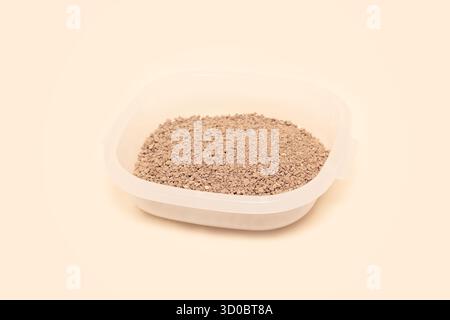 Granuli marroni di lettiera di gatto esposti in un contenitore trasparente su una superficie beige chiaro, evidenziando la consistenza e il colore fini. Cura degli animali domestici Foto Stock
