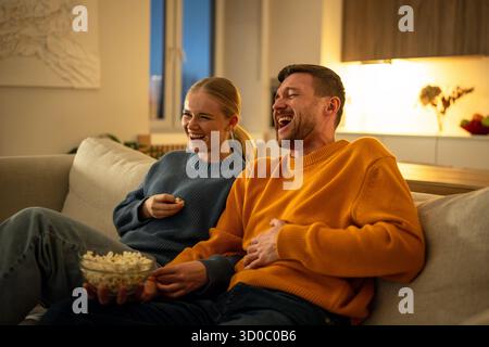 Serata con film in famiglia, giovani coppie che ridono e mangiano popcorn mentre guardano la commedia in una casa accogliente Foto Stock