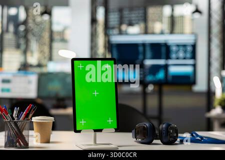 Scrivania da ufficio moderna vuota con tablet che visualizza un modello di schermo verde. Dispositivo digitale sul tavolo nel luogo di lavoro vuoto con spazio di testo isolato per contenuti finanziari o branding aziendale. Foto Stock