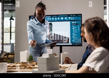 Ingegnere immobiliare afro-americano che presenta il piano di preparazione del sito e le normative sulla suddivisione in zone sui modelli 3D durante una sessione di collaborazione con i colleghi. Futura iniziativa abitativa per la pianificazione urbana. Foto Stock