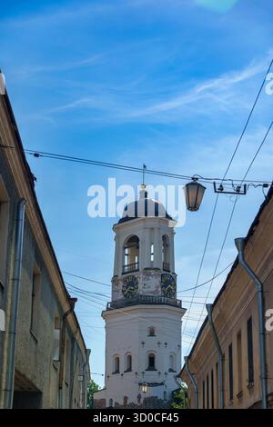 Vista verticale di una vecchia strada europea con pavimentazione in pietra, fiancheggiata da edifici antichi. Una torre dell'orologio bianca sorge sullo sfondo sotto la luminosa S Foto Stock