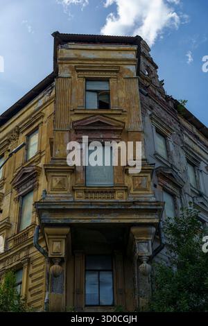 Vista ad angolo basso di un vecchio edificio decadente con facciata gialla sbiadita, elementi architettonici classici e finestre rotte. Cielo nuvoloso sullo sfondo Foto Stock