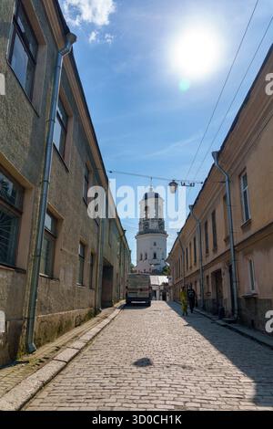 Vista verticale di una vecchia strada europea con pavimentazione in pietra, fiancheggiata da edifici antichi. Una torre dell'orologio bianca sorge sullo sfondo sotto la luminosa S Foto Stock
