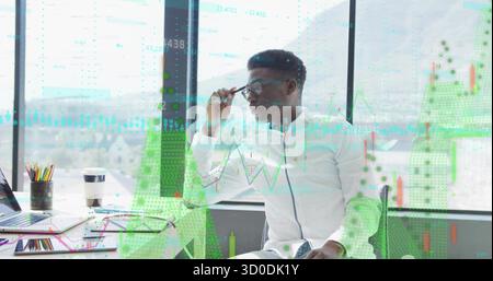 Occhiali regolabili uomo afroamericano che guarda il laptop in ufficio, con matite colorate e grafici Foto Stock