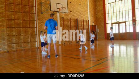 Allenatore con palla arancione che guida i giocatori dribbling nella palestra scolastica, con cornici per arrampicata Foto Stock