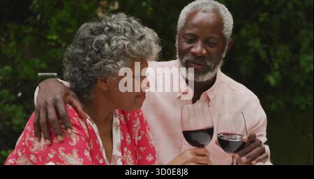 Brinda una coppia senior che solleva bicchieri da vino rosso sul patio del giardino, con fitte foglie verdi Foto Stock