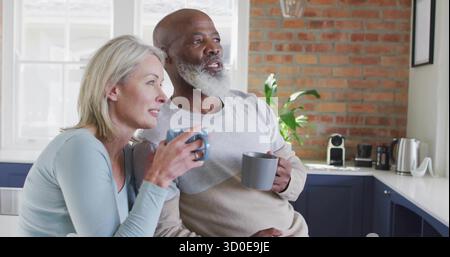 Una coppia anziana che si appoggia al bancone della cucina che guarda fuori dalla finestra, tenendo le tazze in ceramica Foto Stock