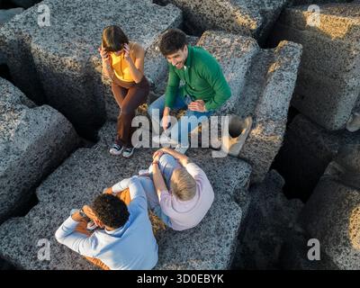 Un drone aereo cattura un gruppo variegato di quattro giovani amici seduti e rilassati insieme su grandi scogliere in riva al mare blu durante un tour Foto Stock