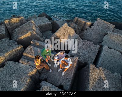 Un drone aereo cattura un gruppo variegato di quattro giovani amici seduti e rilassati insieme su grandi scogliere in riva al mare blu durante un tour Foto Stock