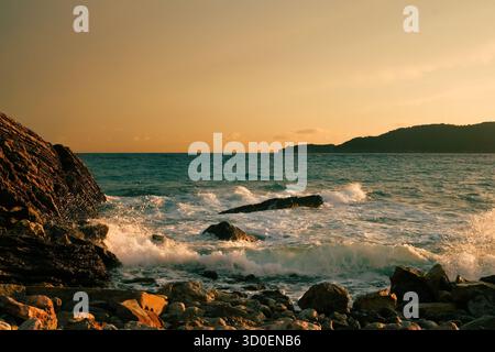 Costa rocciosa con onde che si infrangono contro la costa al tramonto, bagnata dalla luce dorata con colline lontane all'orizzonte Foto Stock