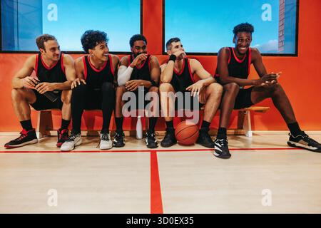 Giovani giocatori di basket maschili seduti su una panchina insieme, sorridenti e chiacchierati, che indicano il lavoro di squadra, l'amicizia e il cameratismo in un luminoso co indoor Foto Stock