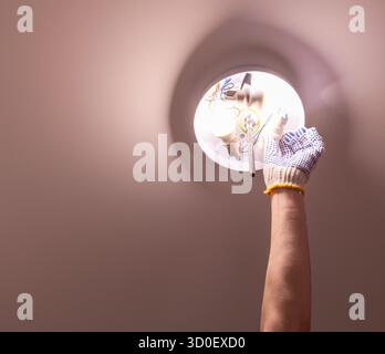 Primo piano di una mano in un guanto di protezione che esegue la manutenzione e l'installazione elettrica a casa. Elettricista che sostituisce una lampadina in un fissaggio a soffitto Foto Stock