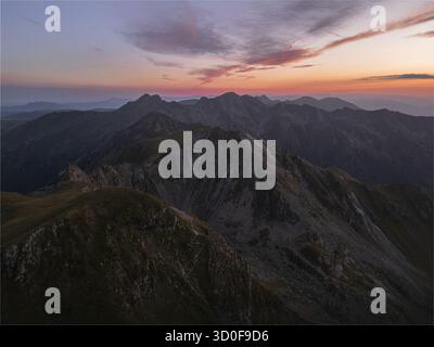 La vista aerea delle aspre cime delle montagne perfora il cielo mentre il tramonto dipinge l'orizzonte con sfumature ardenti, creando un panorama mozzafiato, la Romania. Foto Stock