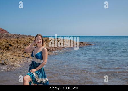 Allegra giovane donna in piedi graziosamente sul bordo del mare durante una soleggiata giornata estiva, adorabile signora che indossa un prendisole a motivi azzurri in posa Foto Stock