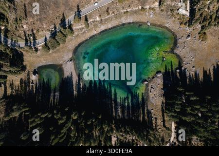 Veduta aerea delle acque intensamente turchesi del Lago di Carezza incastonata tra conifere verde scuro e fogliame autunnale soleggiato, Karersee, T. Foto Stock