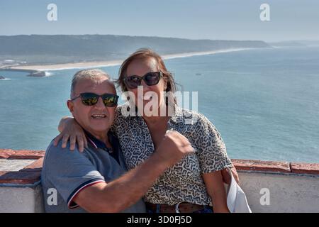 Una coppia più anziana si pone teneramente abbracciando un punto panoramico a Nazare, in Portogallo, con l'Oceano Atlantico e la costa sullo sfondo. Foto Stock