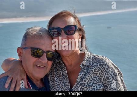 Una coppia più anziana si pone teneramente abbracciando un punto panoramico a Nazare, in Portogallo, con l'Oceano Atlantico e la costa sullo sfondo. Foto Stock