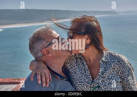 Una coppia più anziana si pone teneramente abbracciando un punto panoramico a Nazare, in Portogallo, con l'Oceano Atlantico e la costa sullo sfondo. Foto Stock