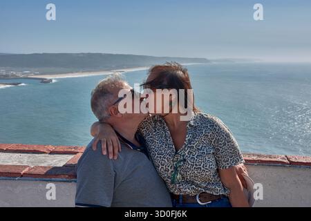 Una coppia più anziana si pone teneramente abbracciando un punto panoramico a Nazare, in Portogallo, con l'Oceano Atlantico e la costa sullo sfondo. Foto Stock