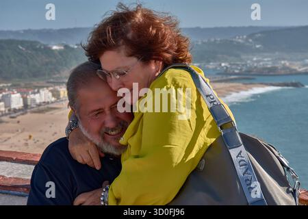Una coppia più anziana si pone teneramente abbracciando un punto panoramico a Nazare, in Portogallo, con l'Oceano Atlantico e la costa sullo sfondo. Foto Stock